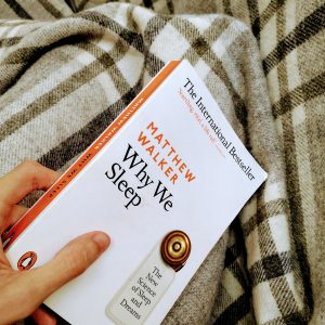 In bed, a hand holds a copy of 'Why We Sleep' by Matthew Walker